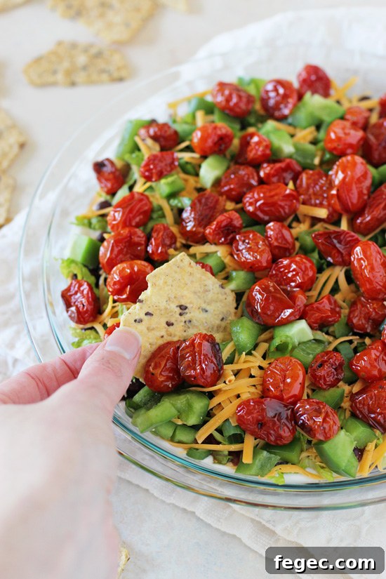 A lighter yet jam-packed with flavor healthier layered taco dip recipe. Colorful, fresh and a crowd-pleasing favorite! A glass pie dish filled with Healthy 7 Layer Dip with a hand scooping dip with a chip.