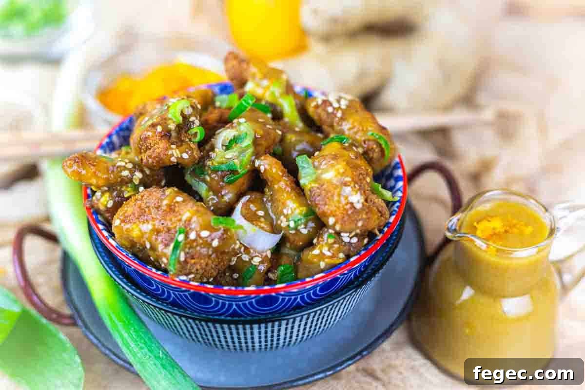 A bowl of fried chicken pieces coated in sauce and garnished with sesame seeds and green onions, accompanied by a small jug of sauce.