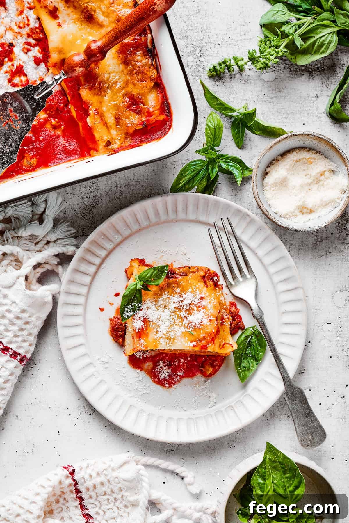 Cheesy Lasagna Roll-Ups Recipe for Family Dinners 3 Overhead view of a lasagna roll up on a plate with a fork, next to a baking dish full of them and some basil leaves.