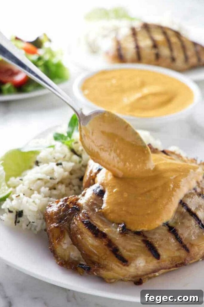 Spoon pours peanut sauce over grilled chicken with rice and lime on plate.