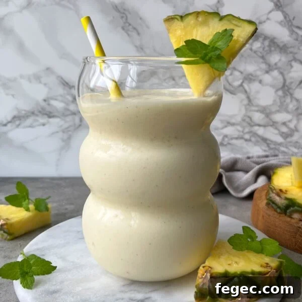 A creamy pineapple smoothie in a curvy glass, garnished with a pineapple slice and mint sprig.