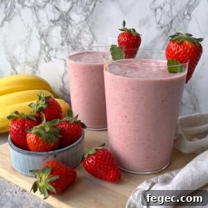 Two glasses of pink strawberry banana smoothie, each garnished with strawberries on the rim, surrounded by fresh strawberries and bananas on a wooden board.