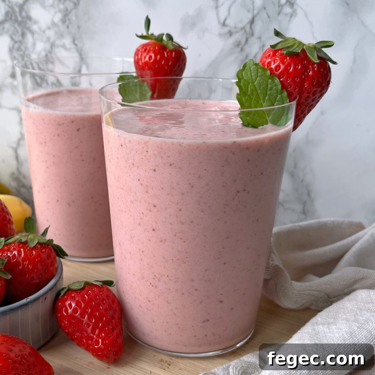 Two glasses of strawberry smoothie garnished with fresh strawberries and mint leaves, set on a wooden surface with extra strawberries and a light-colored cloth nearby.
