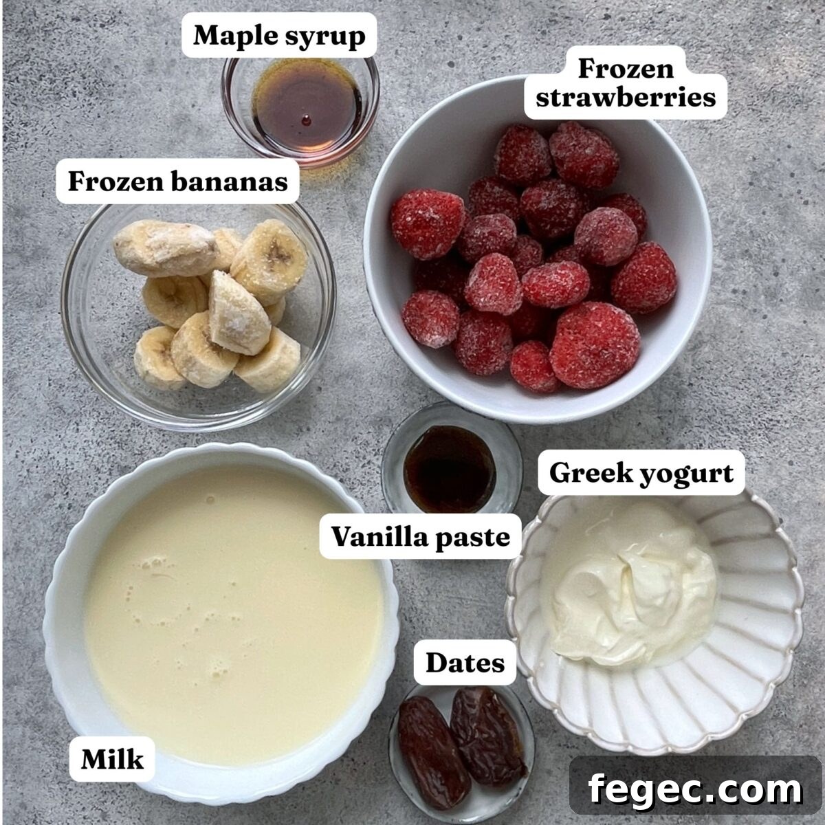 Six labeled bowls and containers display frozen bananas, frozen strawberries, maple syrup, Greek yogurt, dates, vanilla paste, and milk on a gray surface.