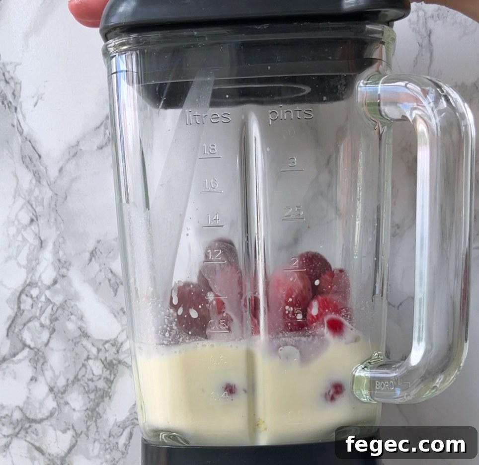 A glass blender contains milk and red strawberries, ready to be blended. The blender has measurement markings in litres and pints.