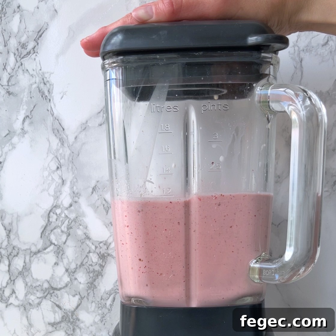A hand holds the lid of a blender containing a pink smoothie, set on a marble countertop.