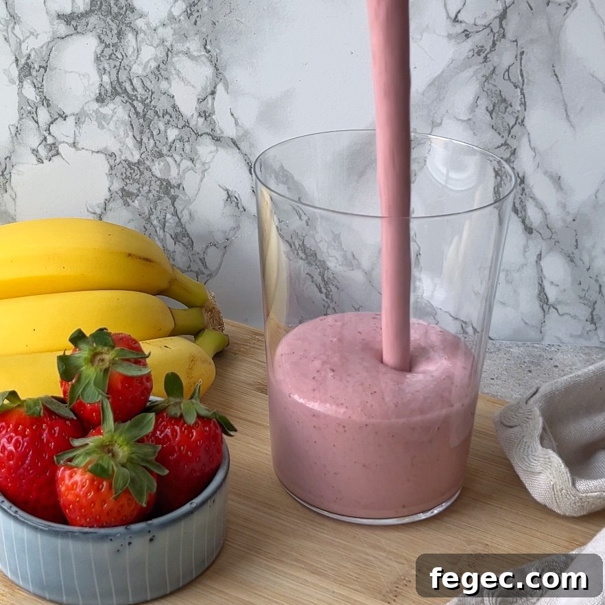 A pink smoothie is being poured into a clear glass, with bananas and strawberries on a wooden surface nearby.
