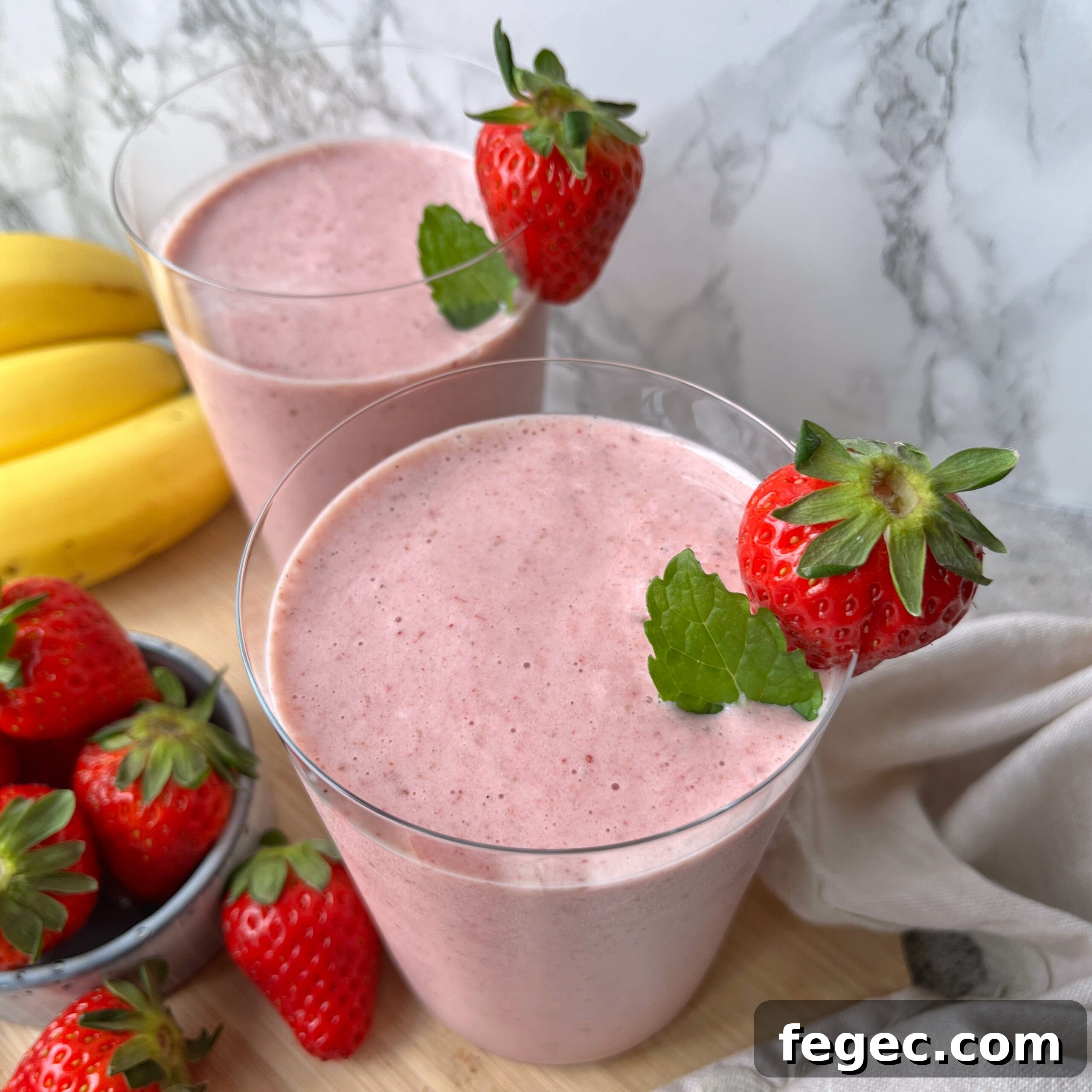 Two glasses of pink strawberry banana smoothie, each garnished with a fresh strawberry and mint leaf.
