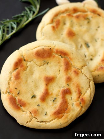 2 Rosemary and Garlic Flatbread rounds sitting on a black table, rosemary and garlic in background.