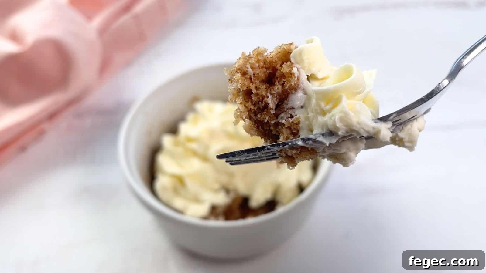 A fork holding a piece of cake with cream cheese frosting over a frosted cake mix mug cake on a table.