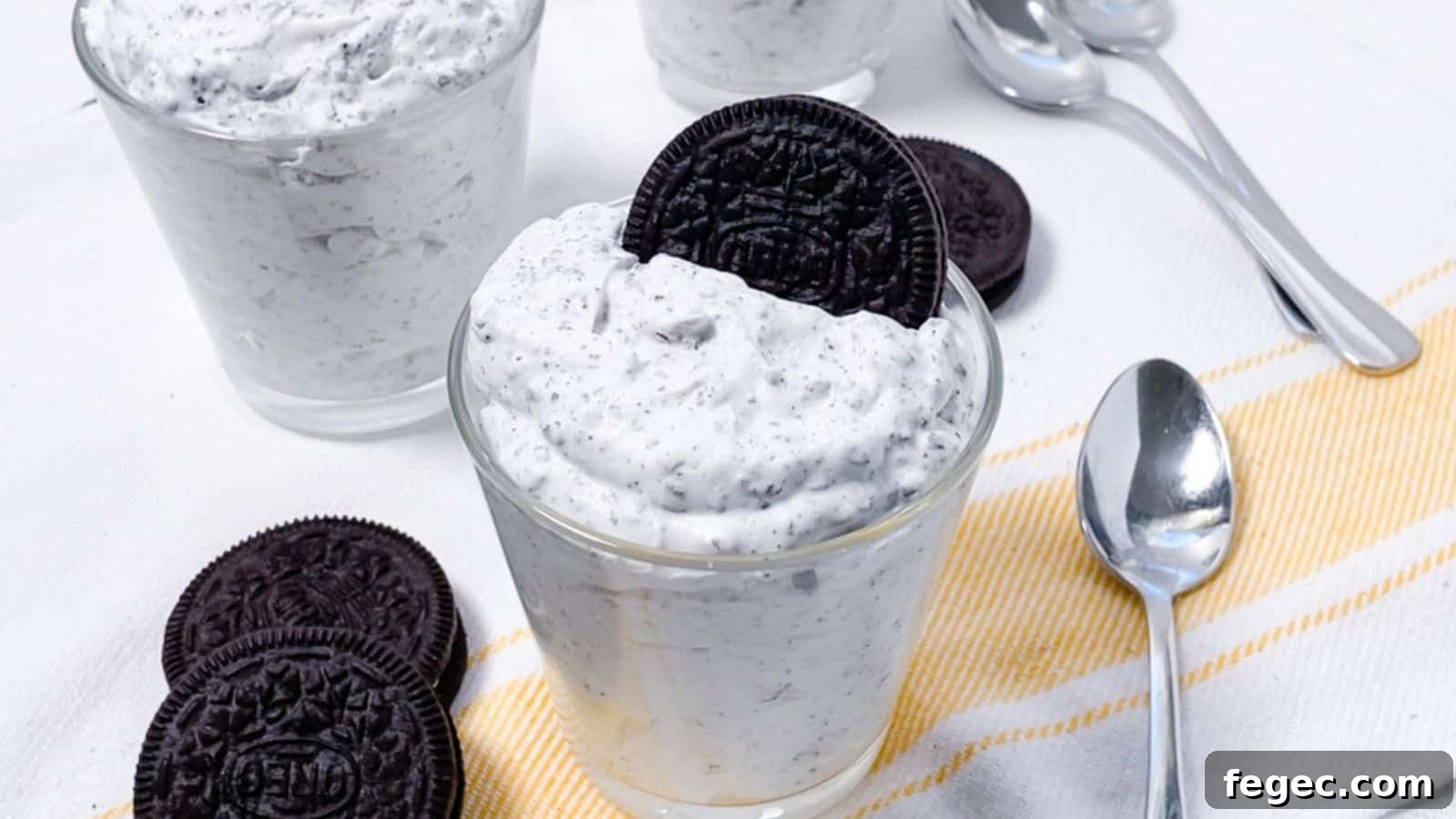 Two glasses of cookies and cream dessert topped with oreo cookies, served on a white tablecloth with a yellow-striped napkin and spoons.