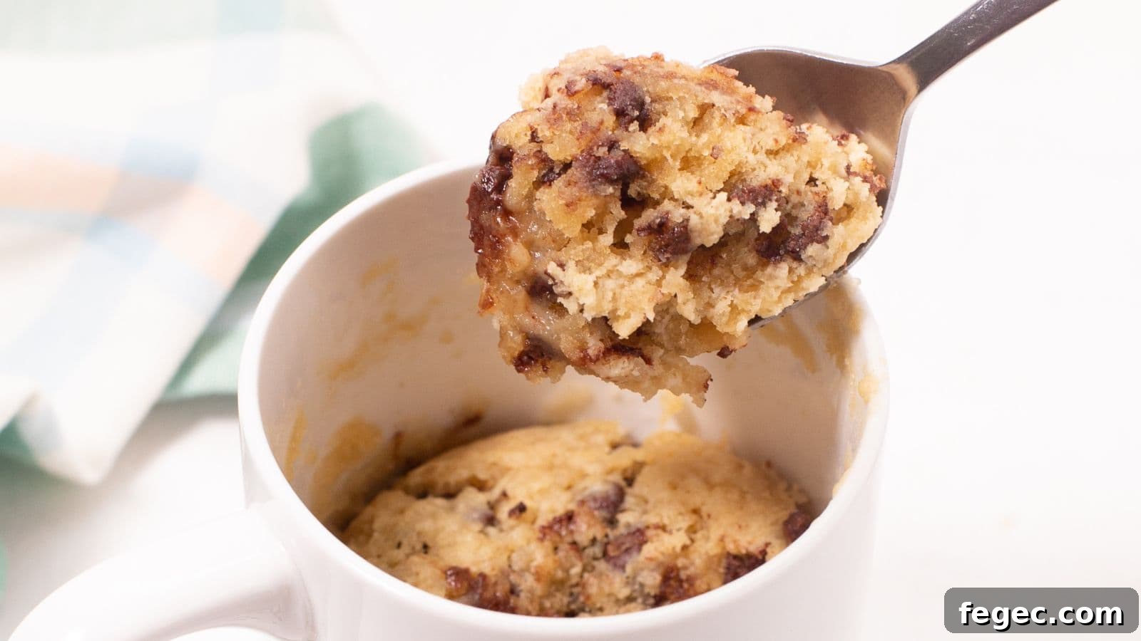 A spoon lifting a bite from a chocolate chip mug cake in a white mug.