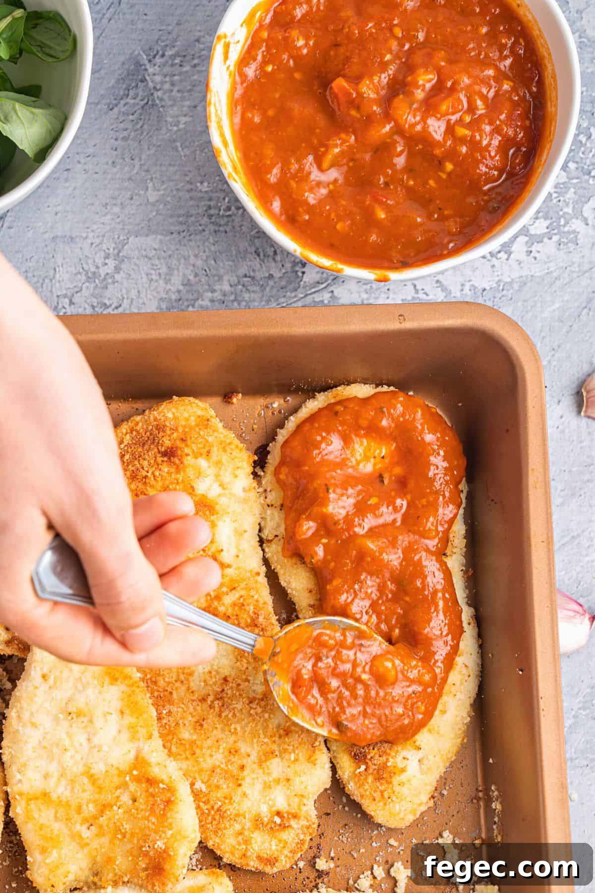 Spreading tomato sauce over breaded chicken cutlets.