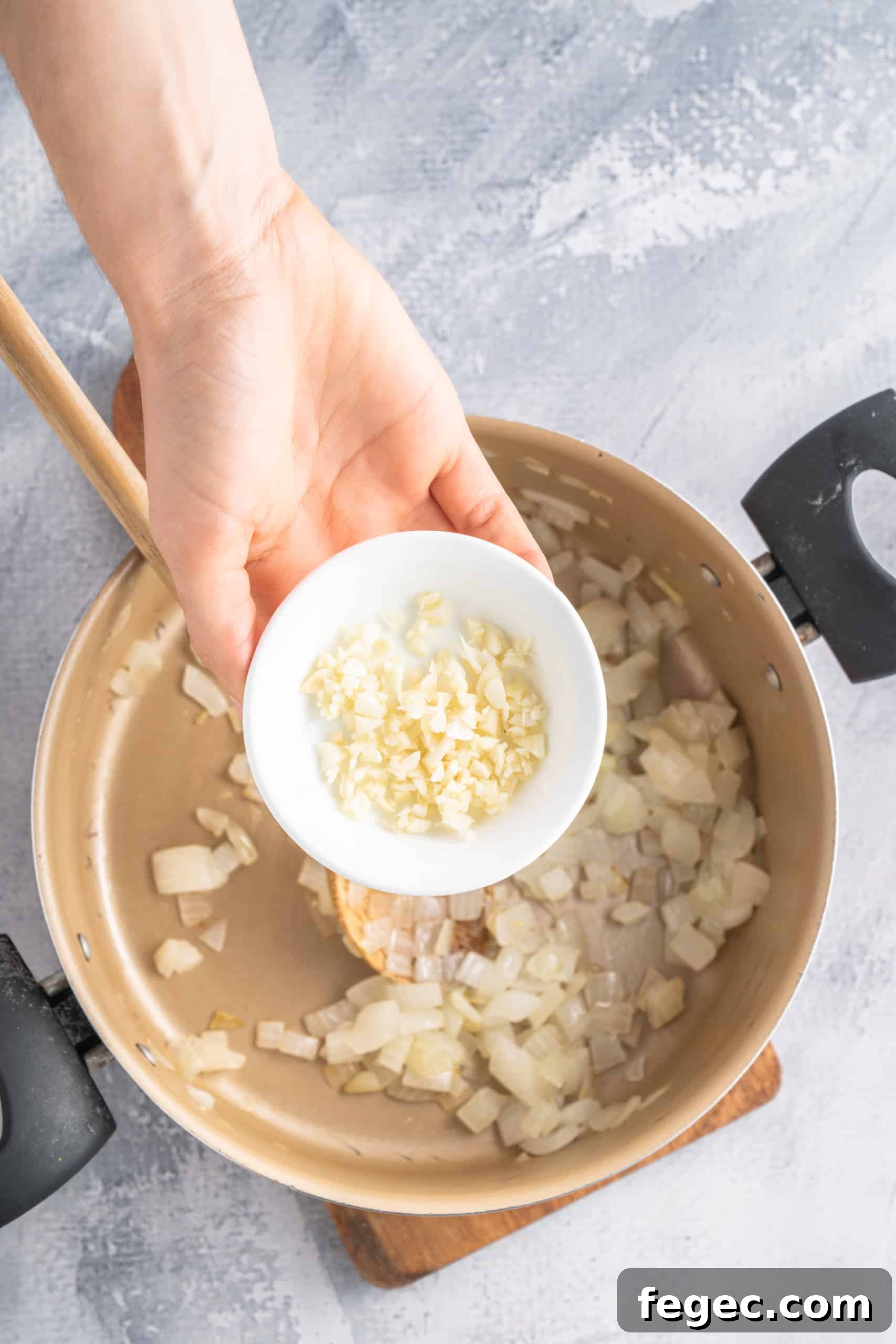 Holding a small bowl of minced garlic over a pot of sautéed onions.