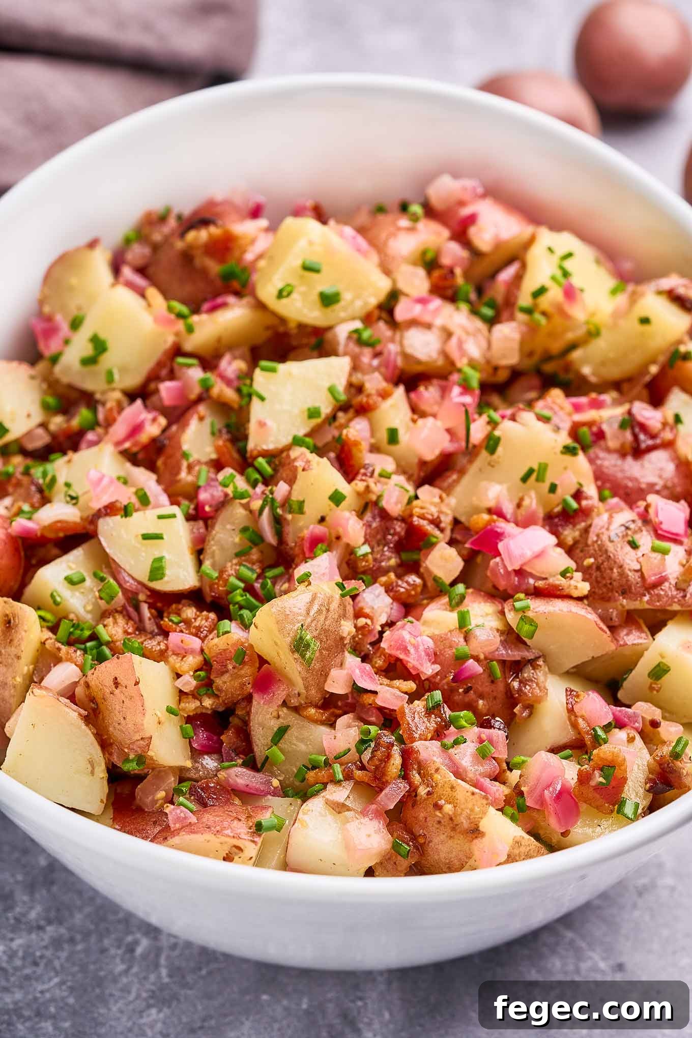 Authentic German Potato Salad with Crispy Bacon and Vinegar Dressing 6 A white oval serving bowl filled with warm German potato salad on a gray concrete surface.