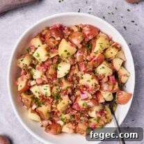 Authentic German Potato Salad with Crispy Bacon and Vinegar Dressing 7 A serving bowl filled with warm potato salad with bacon dressing, serving spoon to the side, surrounded by fresh ingredients and scattered chopped chives.