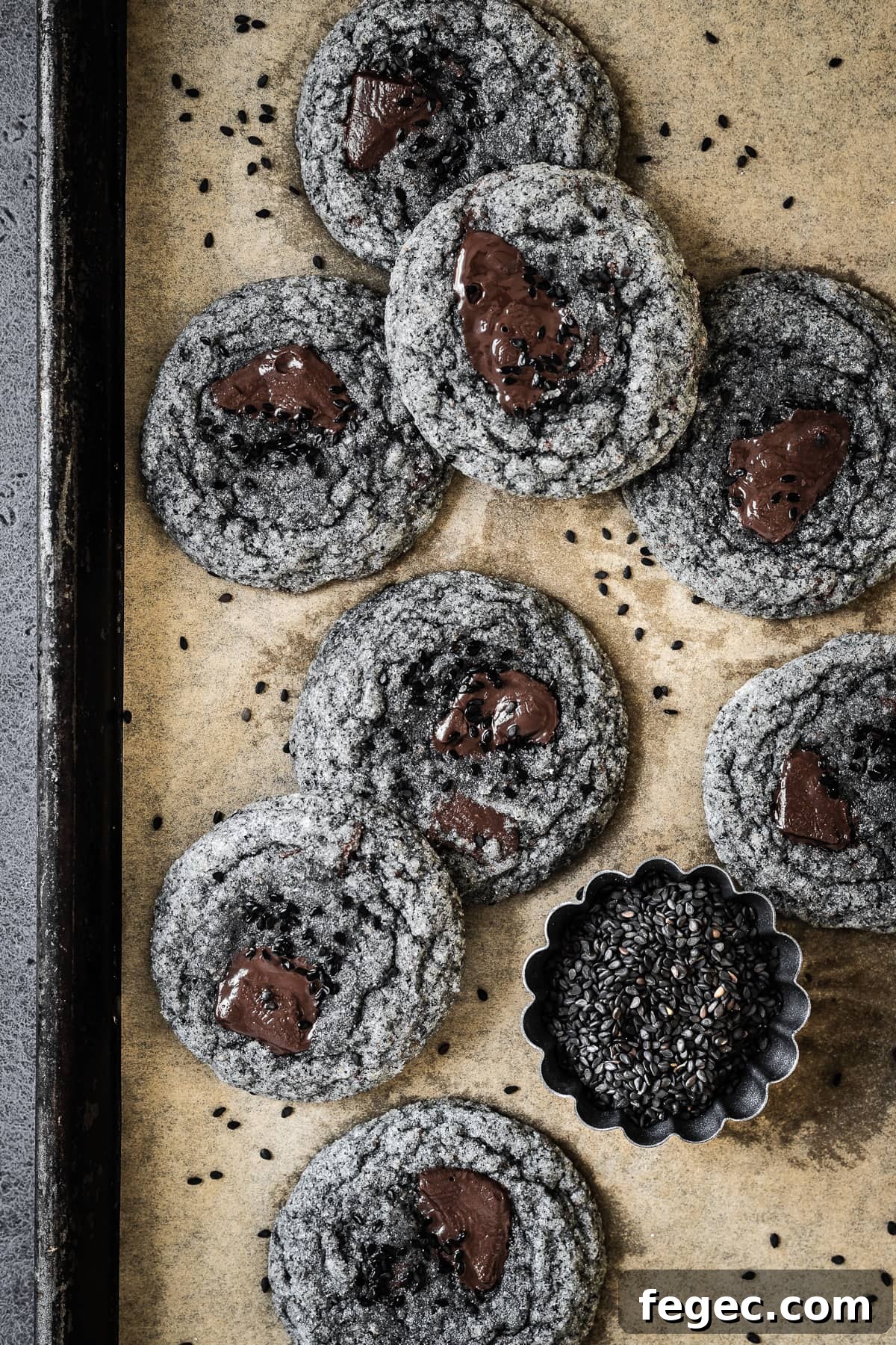Grey cookies with pools of melted chocolate and sesame seeds on a parchment lined baking sheet.