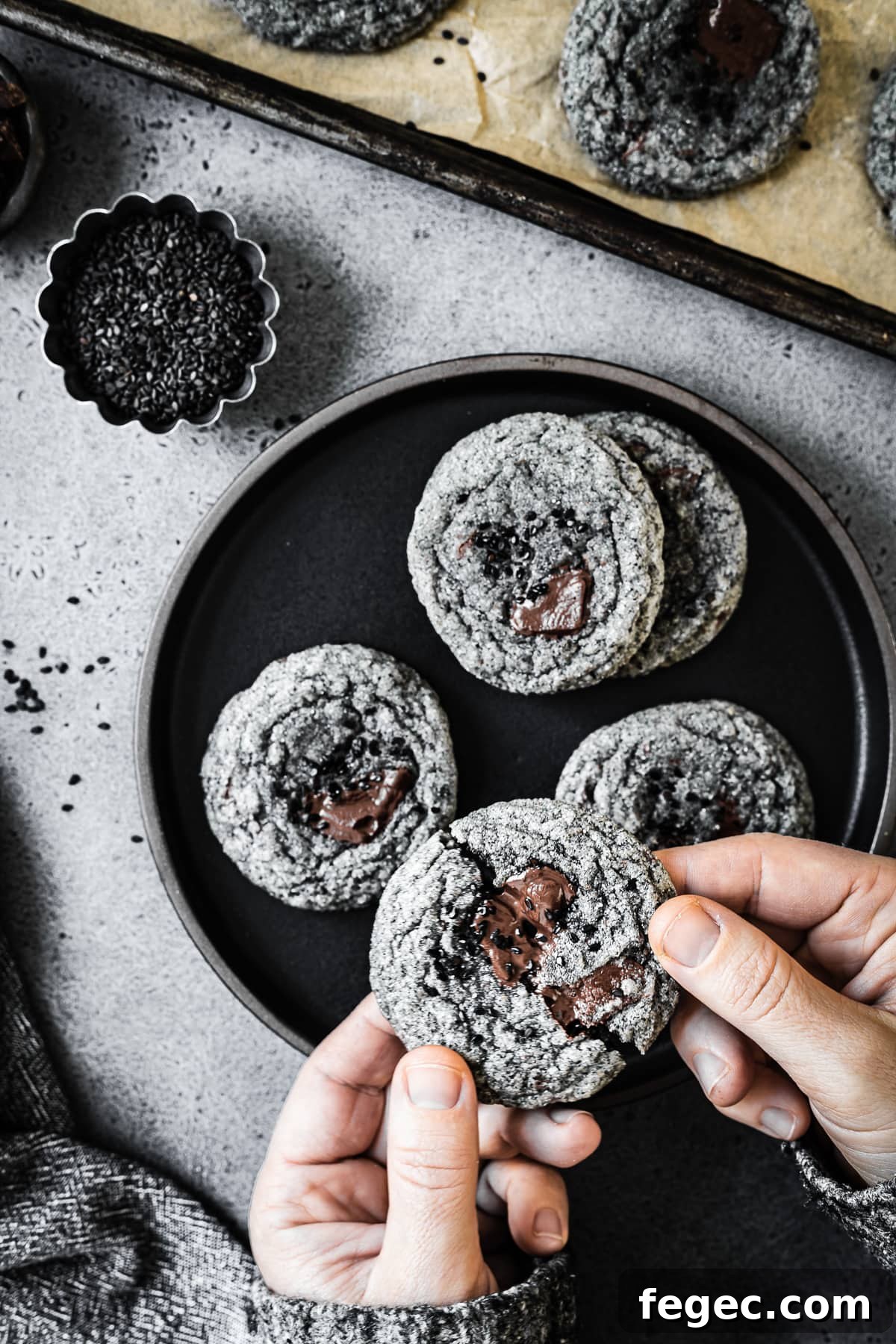 Hands hold a cookie which is being broken in half. There is a black plate full of cookies in the background.