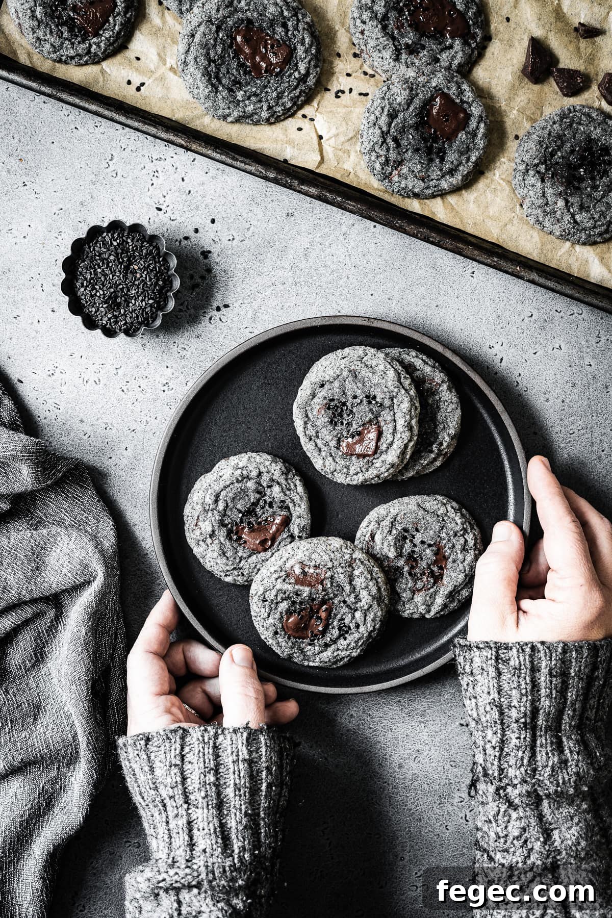 Arms in a grey sweater reach into the frame to hold a black plate of cookies. The colors in the image are monochromatic: grey, black and brown.