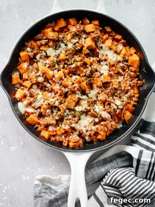 A white skillet filled with a Ground Turkey Sweet Potato Skillet—ground meat, melted cheese, diced sweet potatoes, and herbs—sits on a light gray surface with a striped kitchen towel beside it.