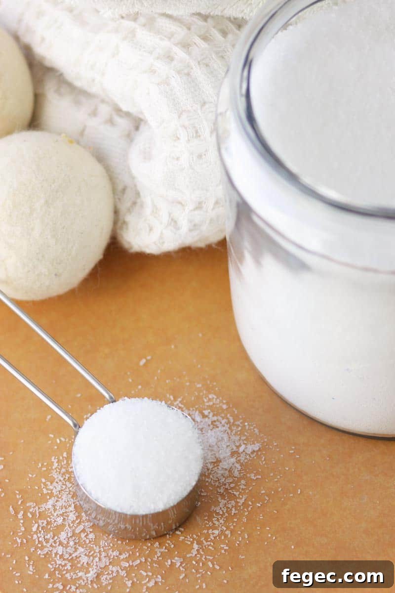 DIY Natural Laundry Scent Booster: Homemade Recipe & Guide 2 a glass jar of scent booster for laundry with a cup measure of the mixture in front of it and a pile of blankets and a dryer ball behind it