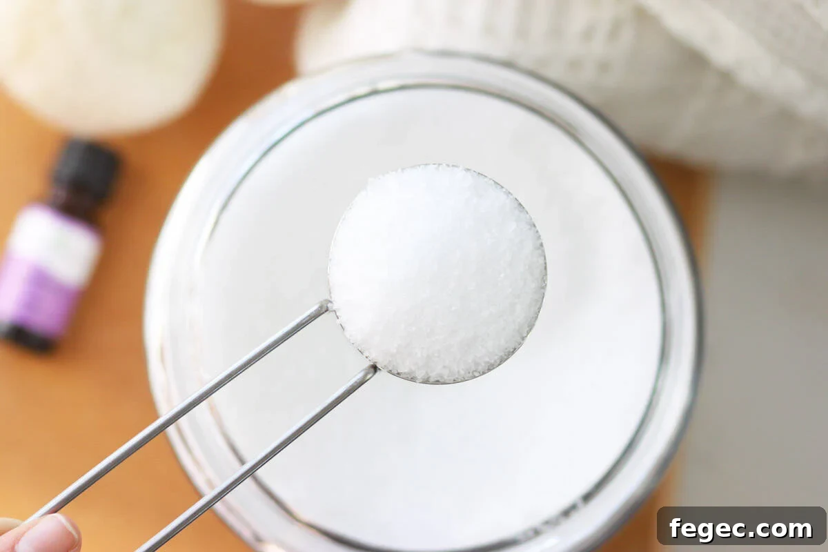 DIY Natural Laundry Scent Booster: Homemade Recipe & Guide 5 a glass jar of scent booster for laundry with a cup measure of the mixture in front of it and a pile of blankets and a dryer ball and essential oil bottles next to it