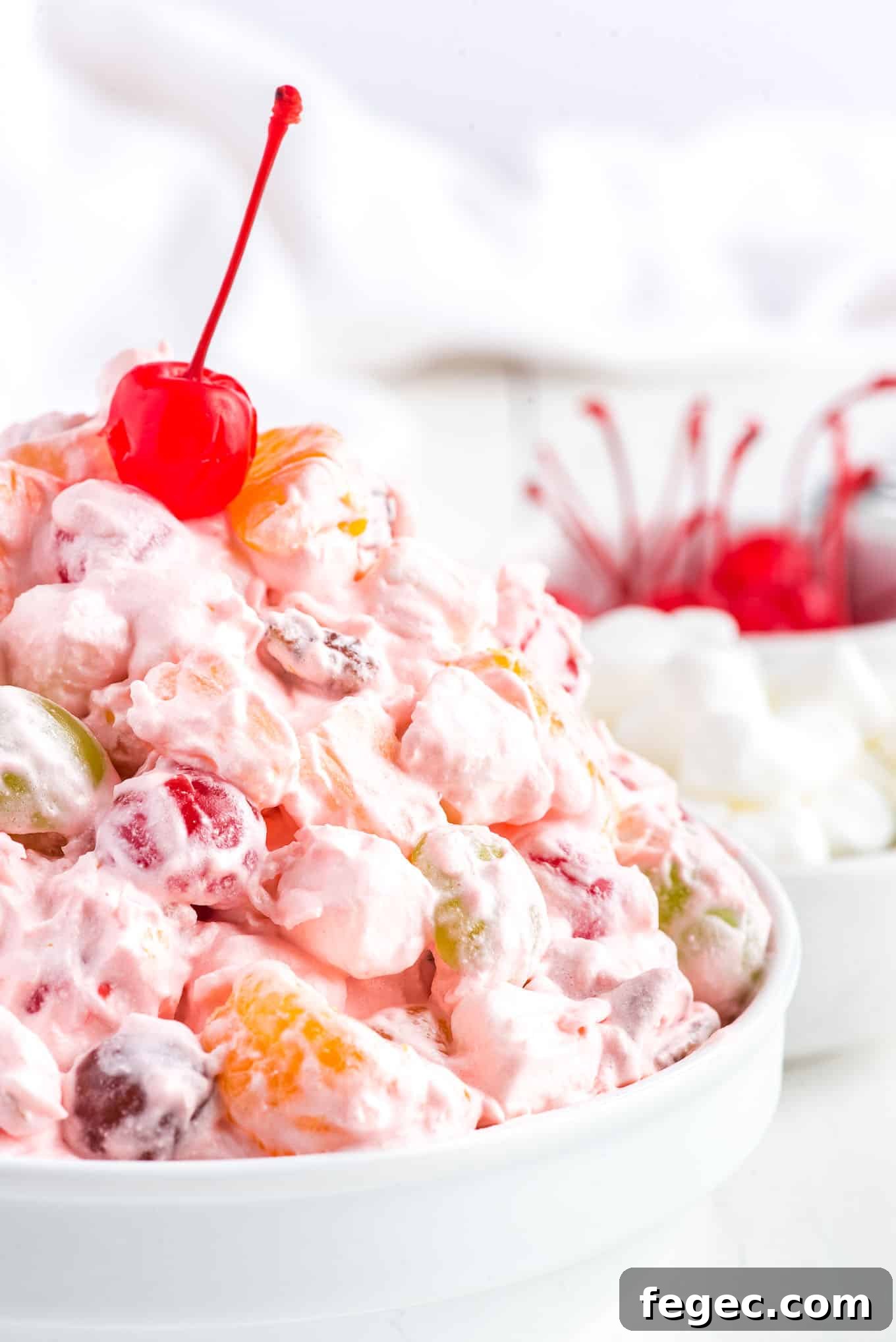 A bowl of fruit salad with Cool Whip on the table in front of a bowl of marshmallows.
