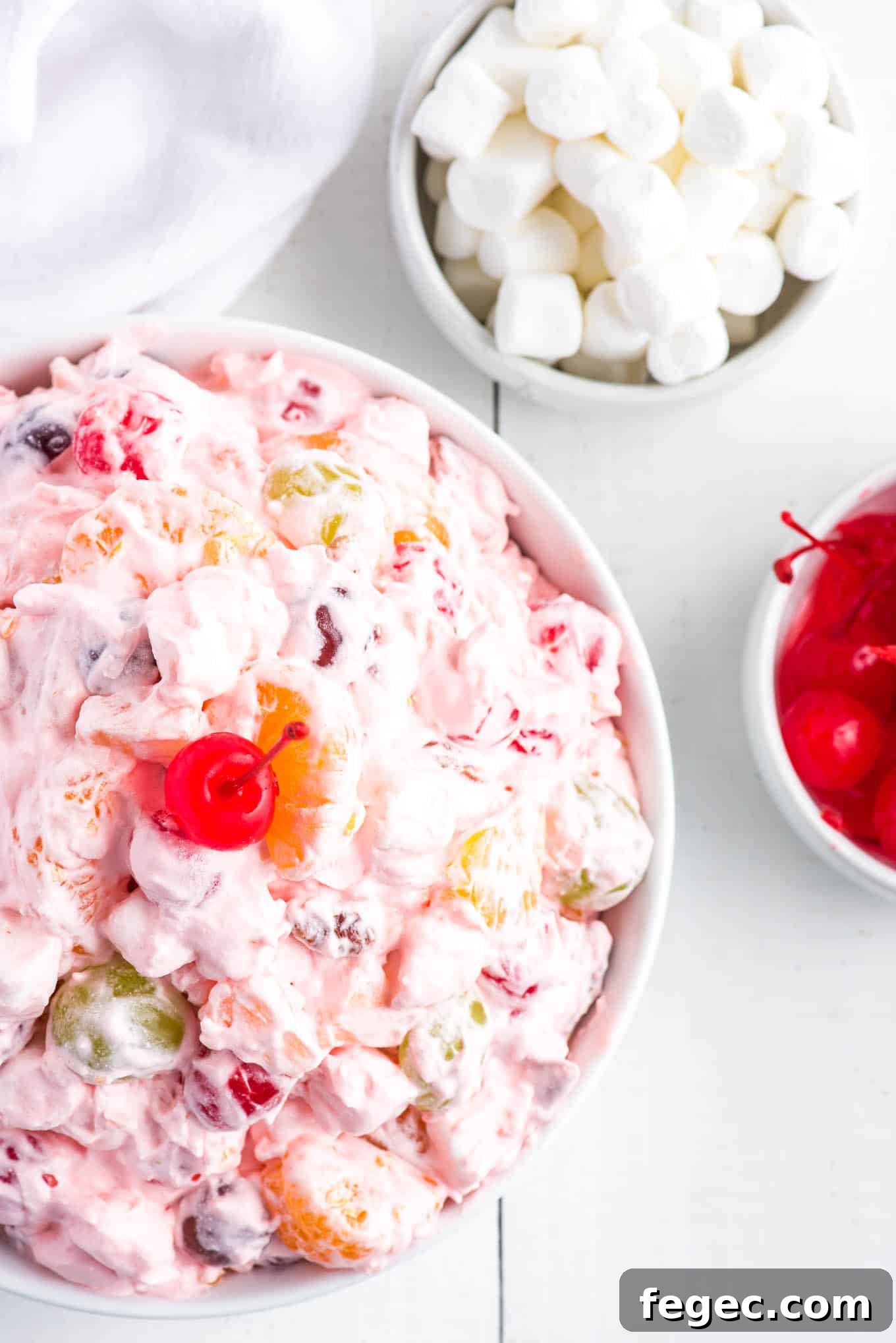 Fruit salad with marshmallows and cool whip on the table with a bowl of marshmallows and cherries.