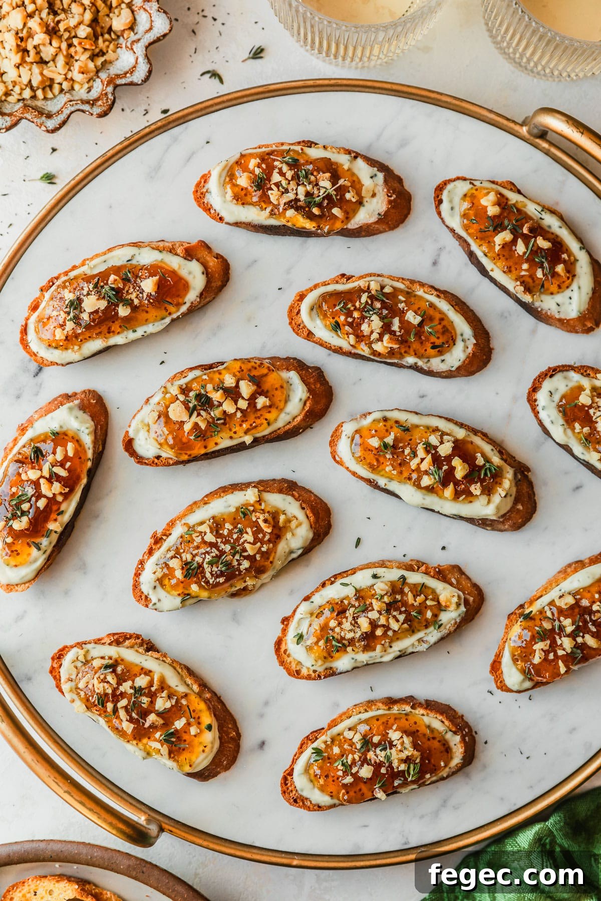 Fig & Goat Cheese Crostini with Honey Drizzle 11 A marble platter with fig, honey, and goat cheese crostini on a white counter next to glasses of white wine, a green linen, and a brown bowl of walnuts.