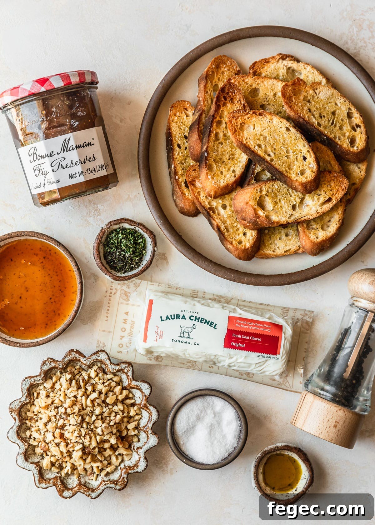 Fig & Goat Cheese Crostini with Honey Drizzle 3 White and brown bowls of bread, jam, honey, herbs, salt, oil, walnuts, and cheese on a tan counter.