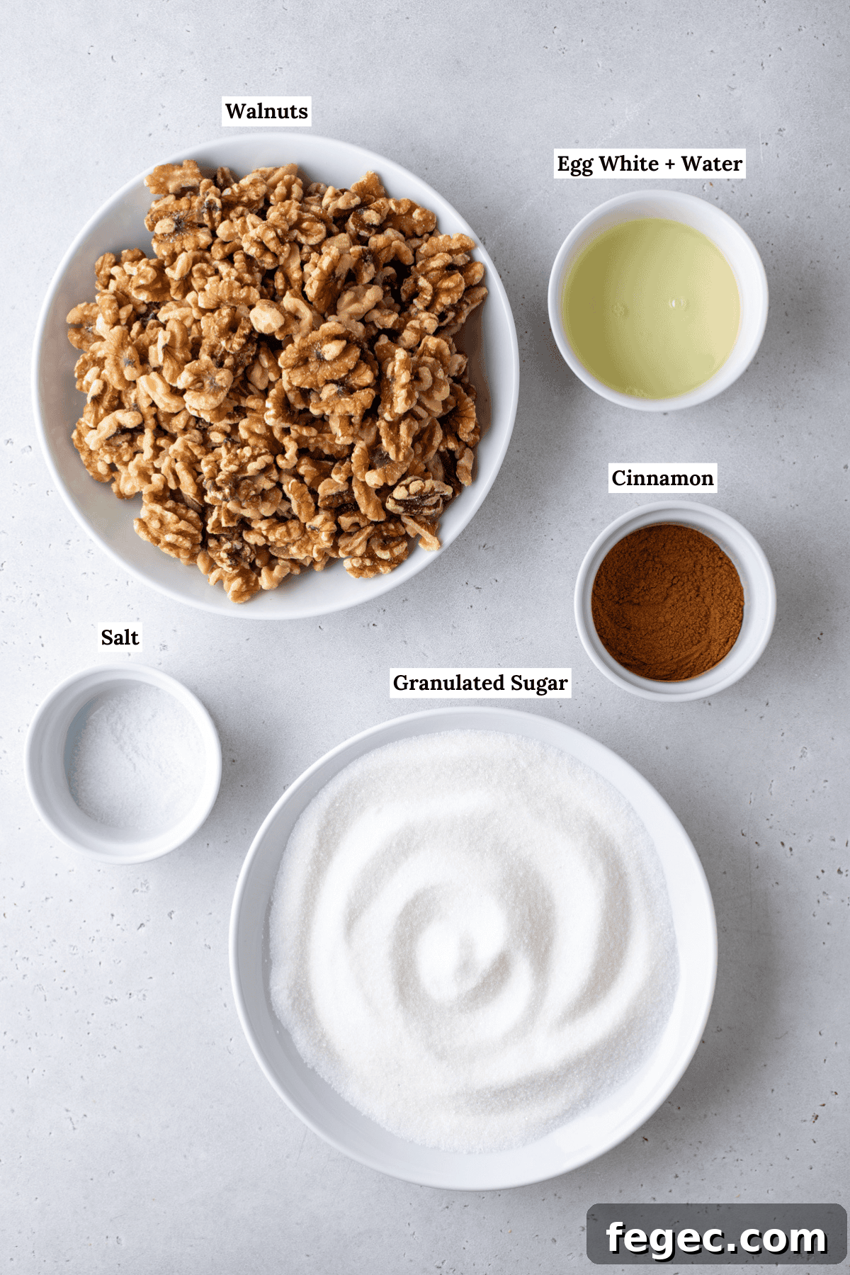 over head view of the ingredients for candied walnuts including walnuts, egg whites, water, salt, cinnamon, and granulated sugar
