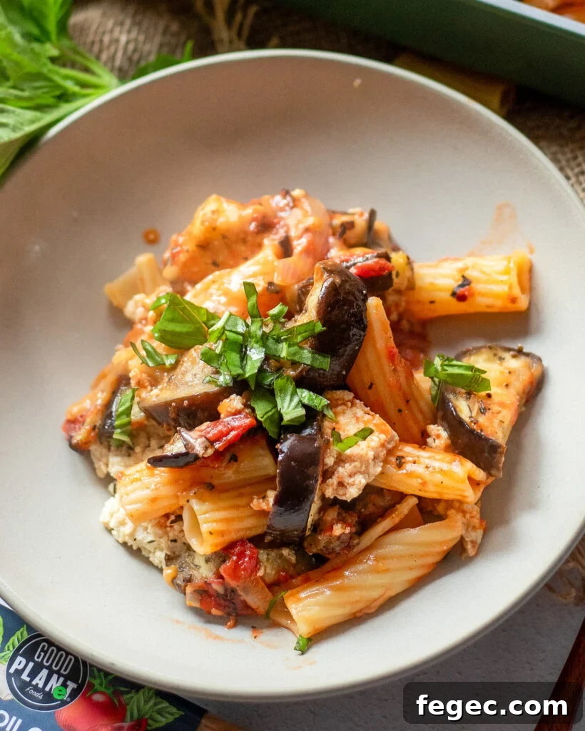 close up view of vegan eggplant pasta in a white dish with roasted eggplant, peppers, tomato sauce, and fresh basil.