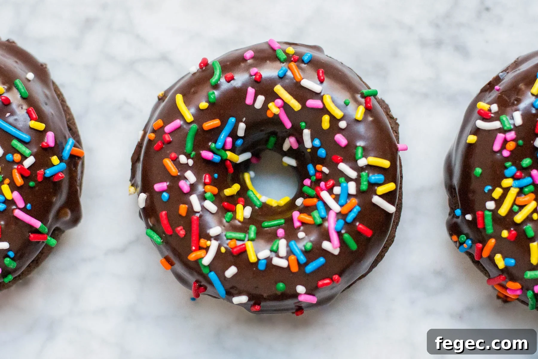 An aerial view of three gluten-free vegan baked chocolate donuts with chocolate glaze and rainbow sprinkles.