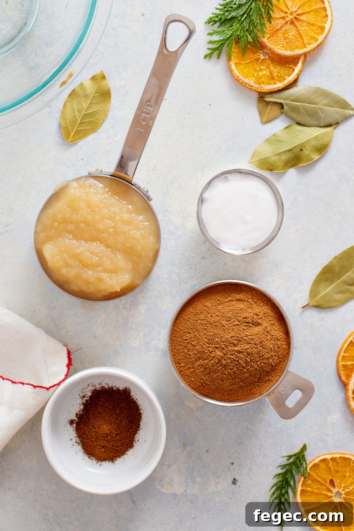 Overhead photo of a flat lay arrangement showing ingredients in metal and ceramic measuring cups: one cup of applesauce, one cup of white liquid glue, one cup of ground cinnamon, and a small bowl of ground cloves. The ingredients are placed on a light surface with scattered dried orange slices, bay leaves, and evergreen sprigs.