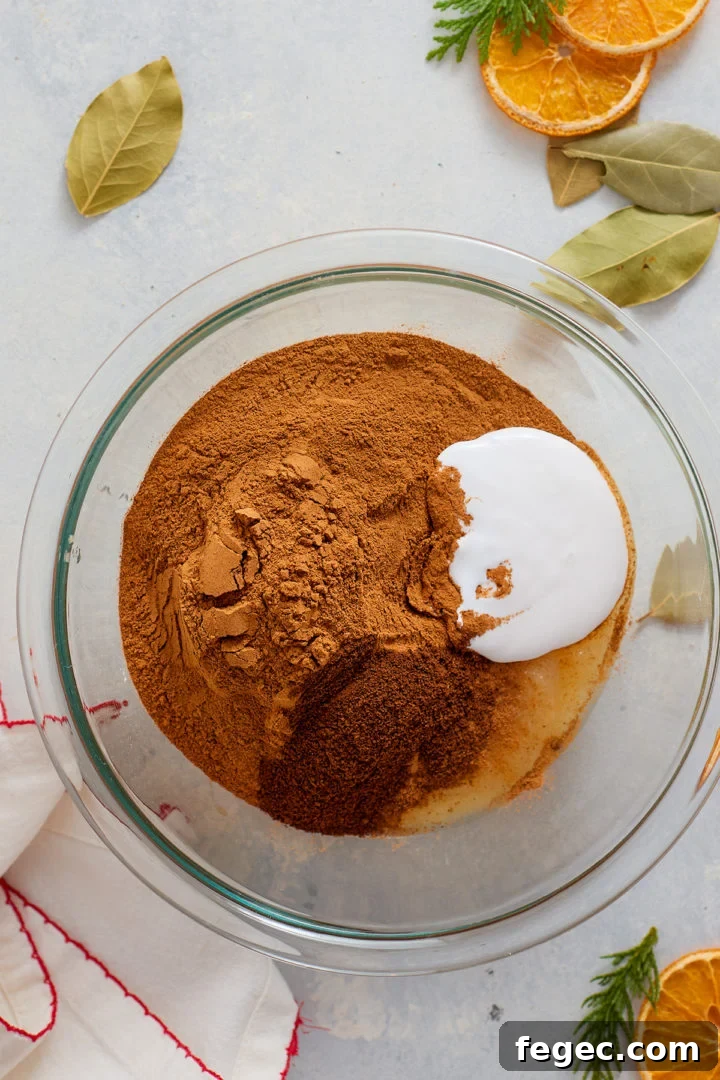 A clear glass mixing bowl contains a mound of unmixed ingredients: ground cinnamon, white liquid glue, and applesauce. The mixture is not yet stirred. Bay leaves, dried orange slices, and evergreen clippings are scattered on the light-colored surface around the bowl.