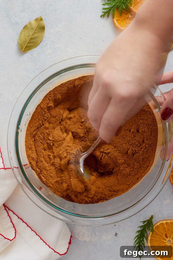 A person’s hand is using a metal spoon to stir a large amount of ground cinnamon into a mixing bowl containing wet ingredients. Only the hand, spoon, and part of the cinnamon mixture are visible. Bay leaves, dried oranges, and greenery decorate the surrounding surface.