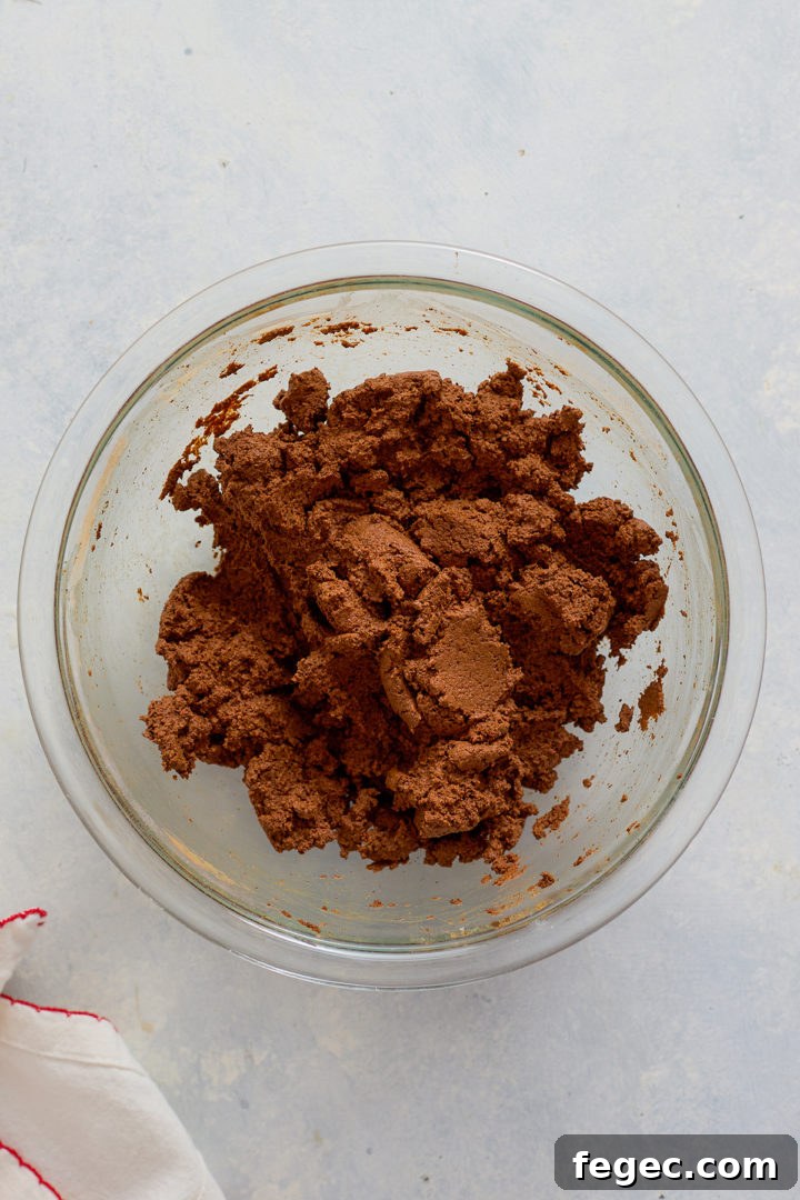 A glass mixing bowl filled with brown cinnamon dough sits on a light surface. The dough appears thick and slightly crumbly, sticking to the sides of the bowl. A white cloth with red stitching is partially visible in the bottom corner.
