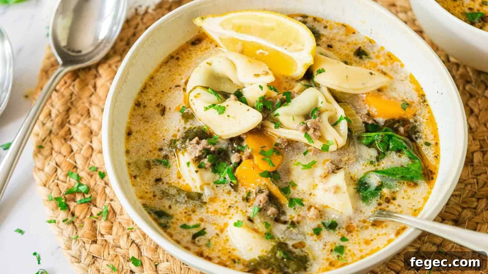 A bowl of creamy tortellini soup with spinach, lemon wedges, and herbs, served with a spoon.