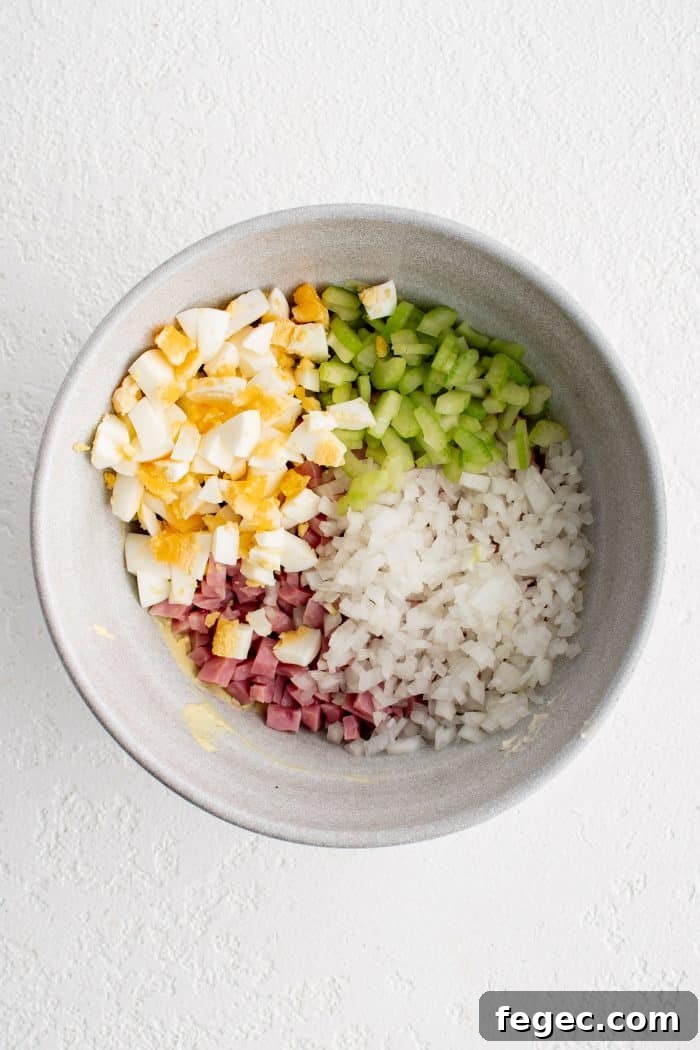 Large white mixing bowl with diced celery, diced onion, diced, ham, and diced hard-boiled egg.