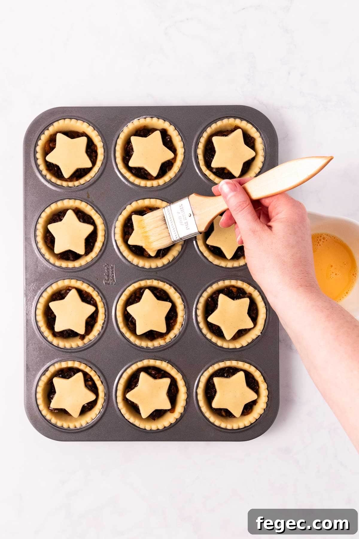 A hand holding a pastry brush, brushing an egg wash on the tops of the mince pies.