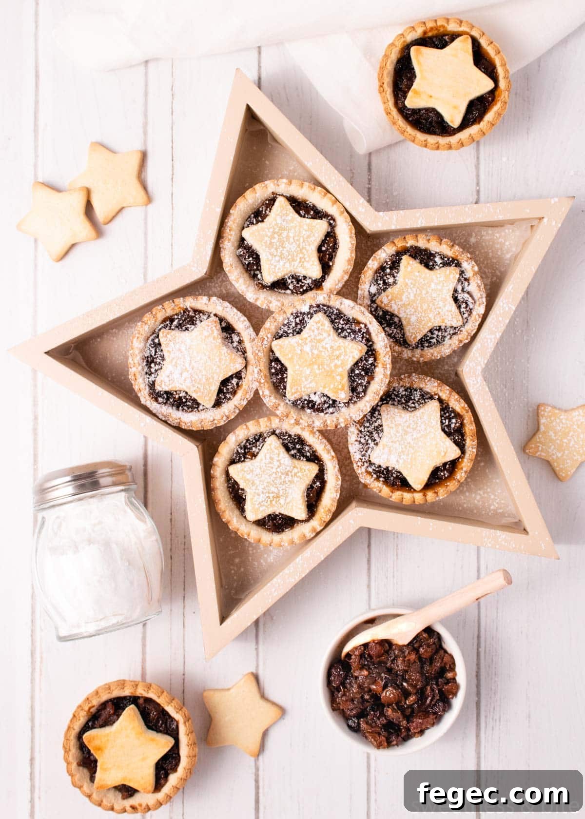 Gluten free mince pies served in a star-shaped wooden box, on a white wooden background, with a small bowl of fruit mince and shaker of icing sugar.