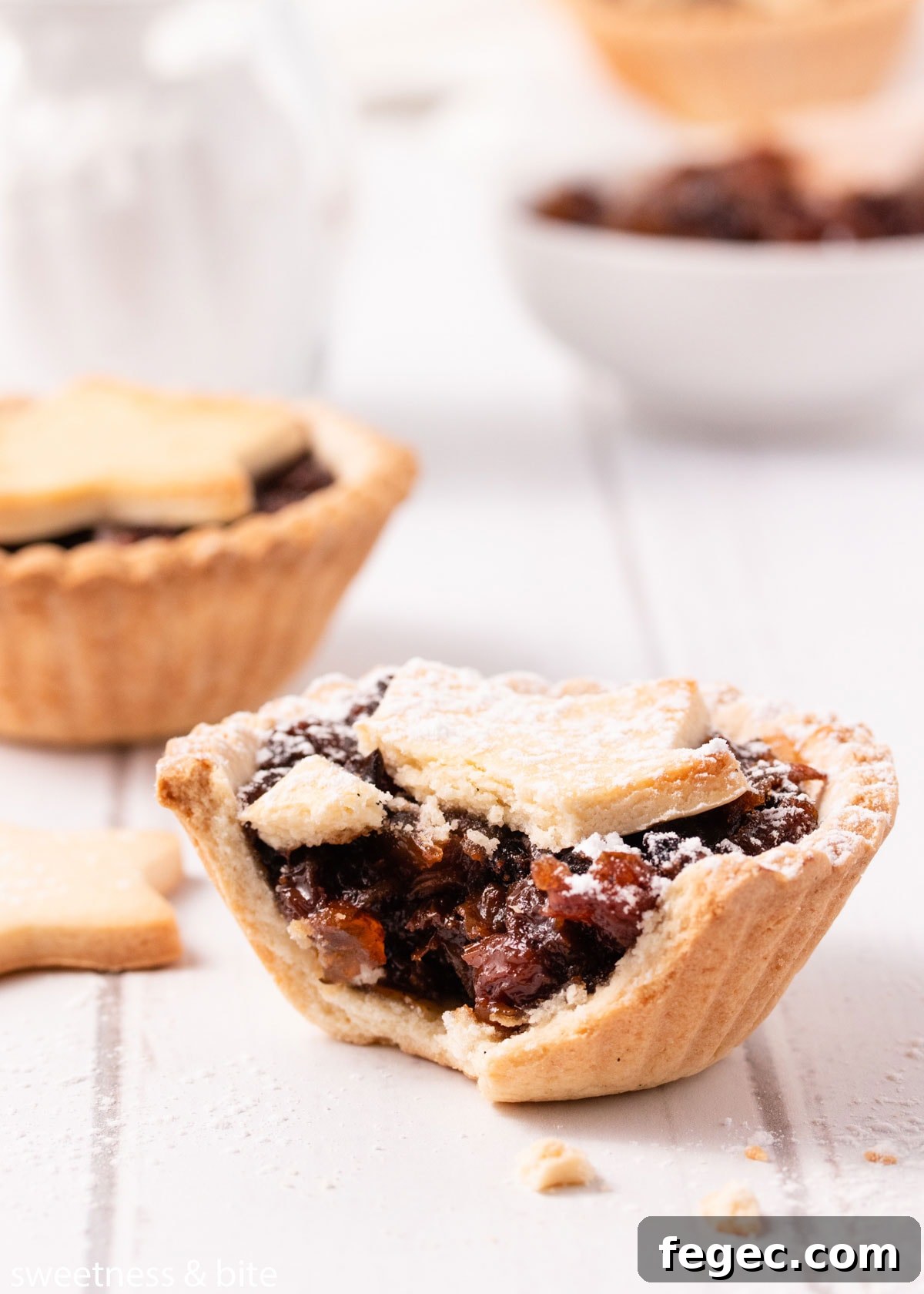 A close up of a bitten mince pie dusted with icing sugar, showing the fruit mince filling and the crunchy biscuit base.