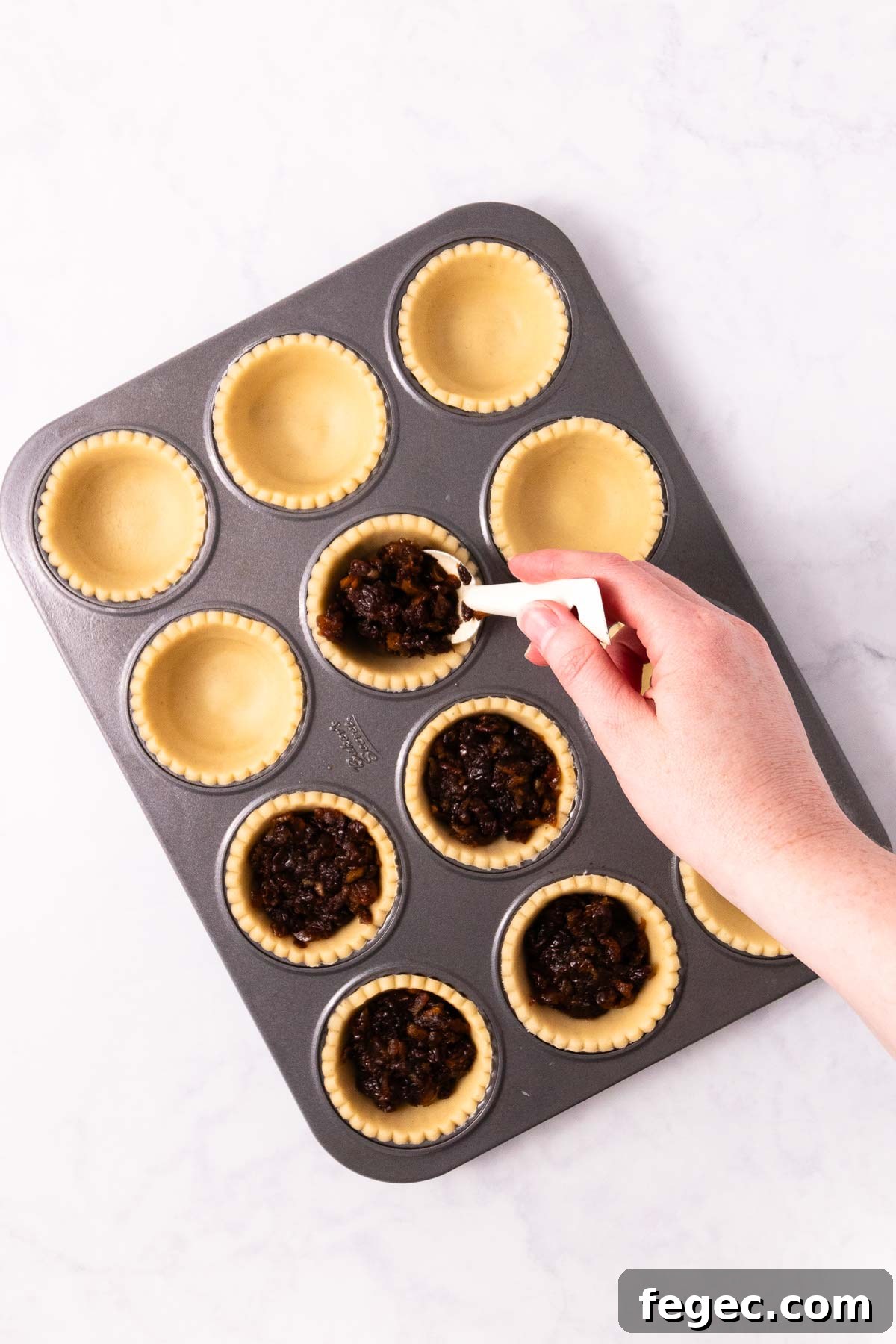 A hand spooning fruit mince into one of the pie cases.