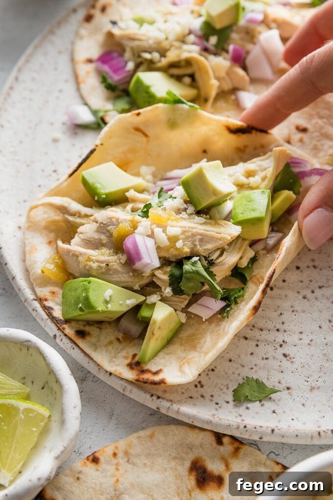Hand holding a salsa verde chicken taco with avocado and diced onion, over a small ceramic plate.