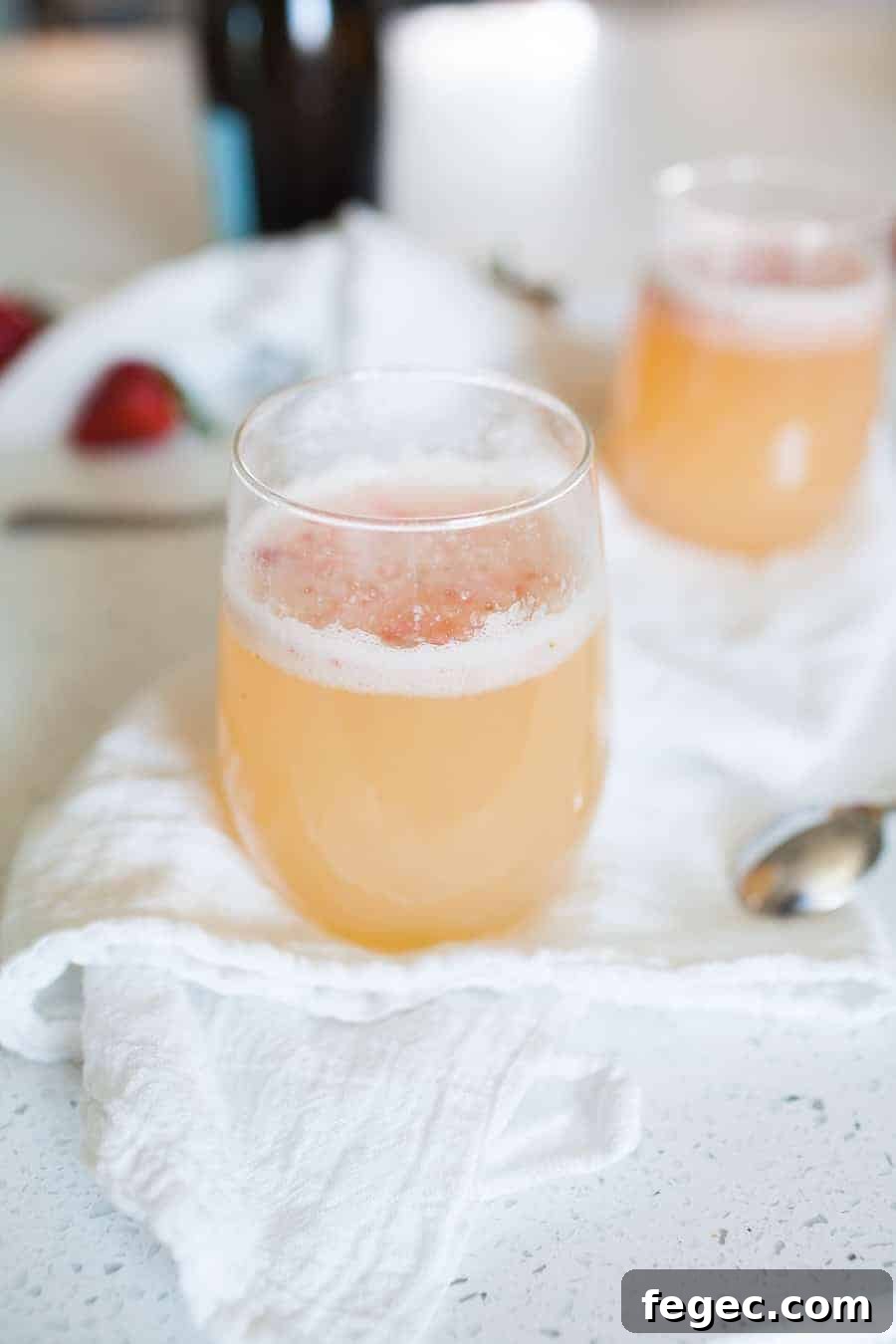 A close-up of a stemless glass filled with a frothy light orange Bellini recipe, placed on a white cloth with a spoon nearby. Another similar drink and strawberries are blurred in the background.