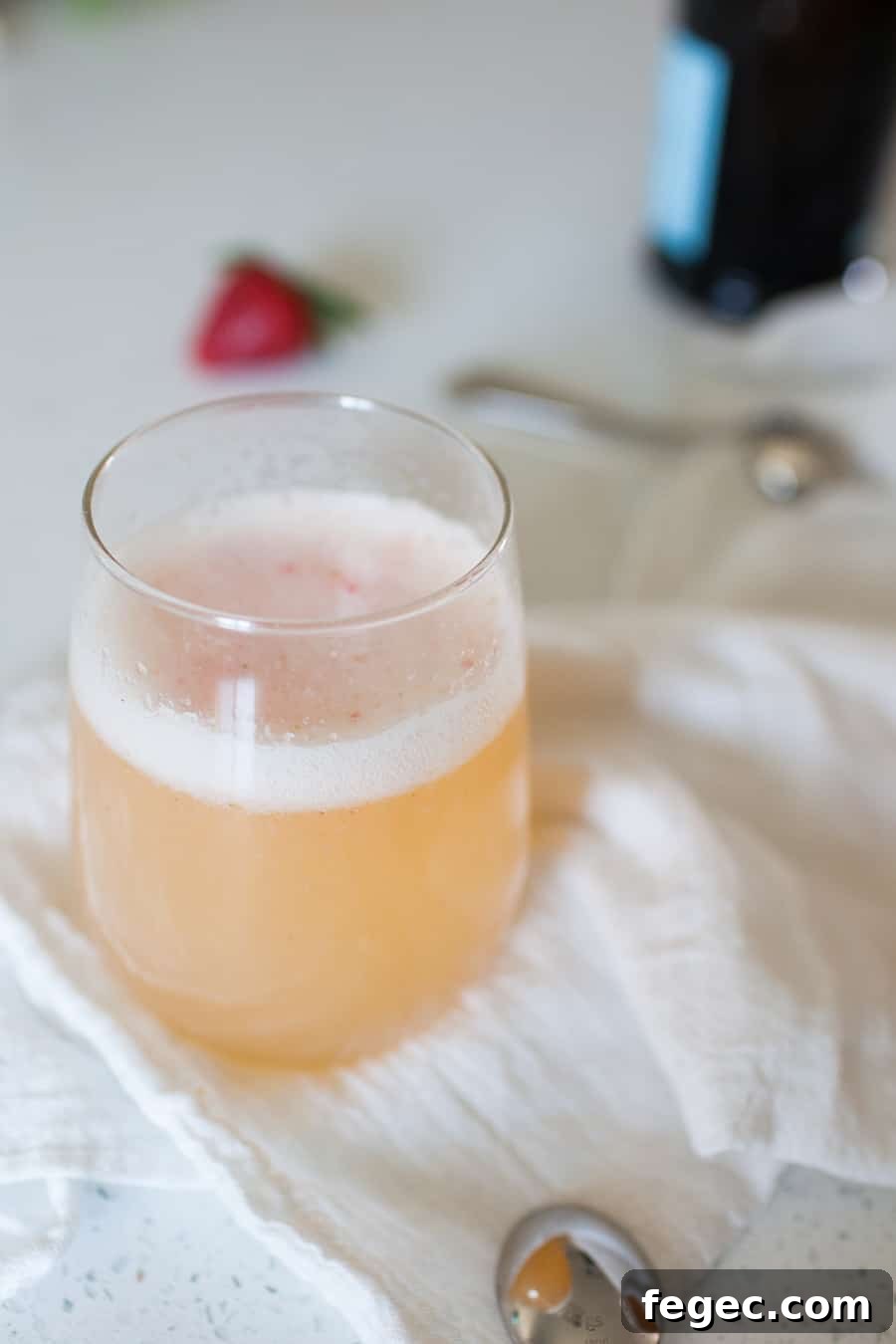 A glass of light pink, frothy Bellini recipe beverage sits on a white cloth with a spoon nearby; a blurred strawberry and bottle are in the background.