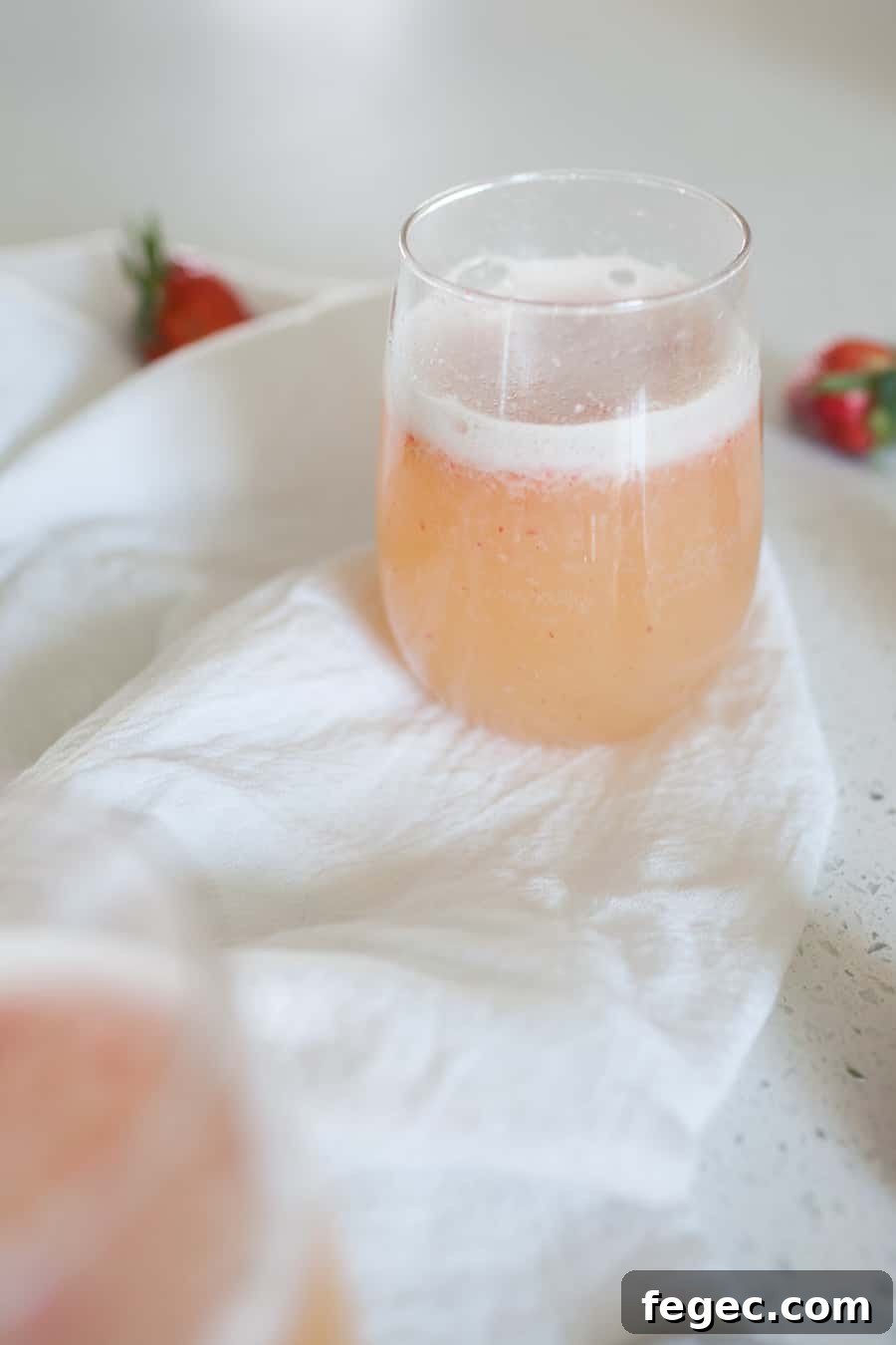 A glass of light pink, frothy beverage sits on a white cloth with blurred strawberries in the background, capturing the refreshing essence of a classic Bellini recipe.
