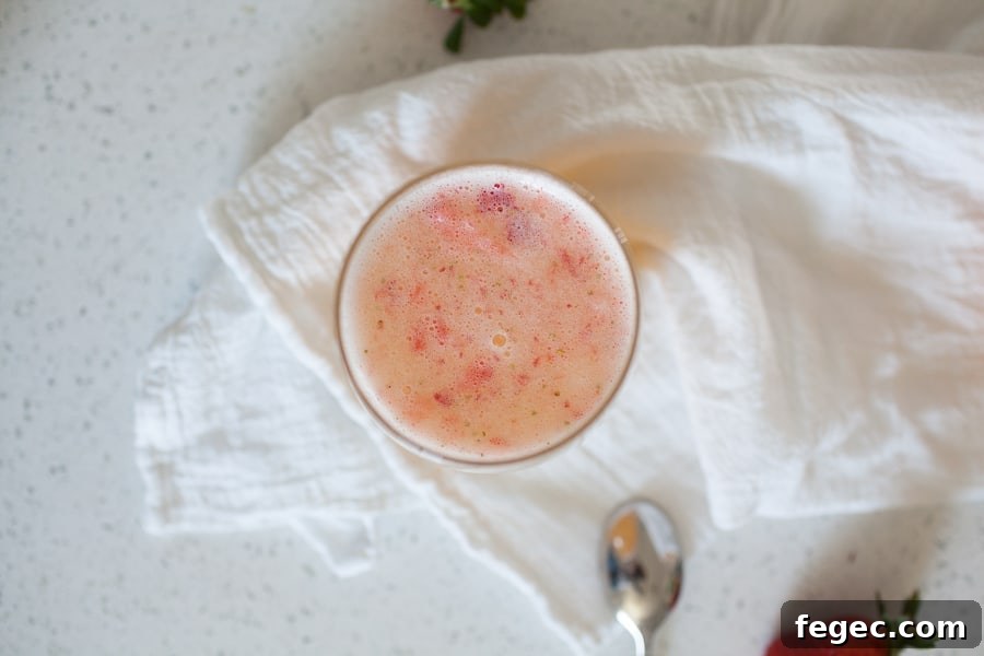 A glass of frothy pink smoothie sits on a white cloth napkin, with a spoon and part of a strawberry nearby, evoking the playful elegance of a classic Bellini recipe.
