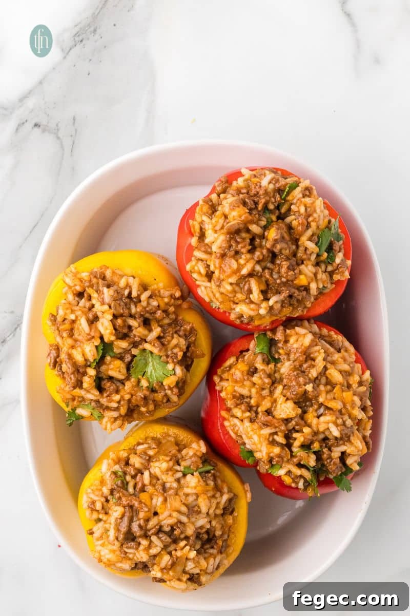 Four bell pepper halves (two red and two yellow) are filled with the ground beef and rice mixture and arranged in a white oval baking dish.