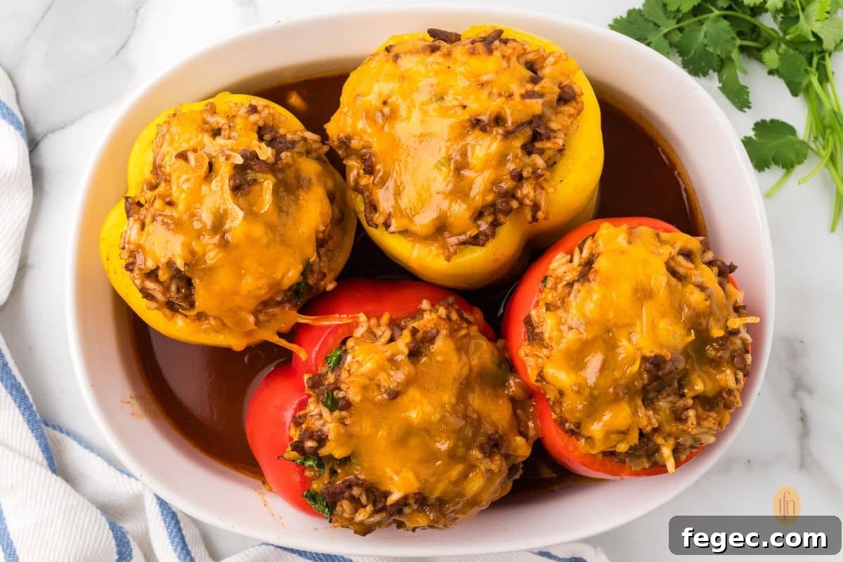 An overhead shot of four baked taco stuffed peppers (two red and two yellow) in a white oval baking dish. The peppers are filled with a cheesy, meat and rice mixture, and are sitting in a rich red sauce.